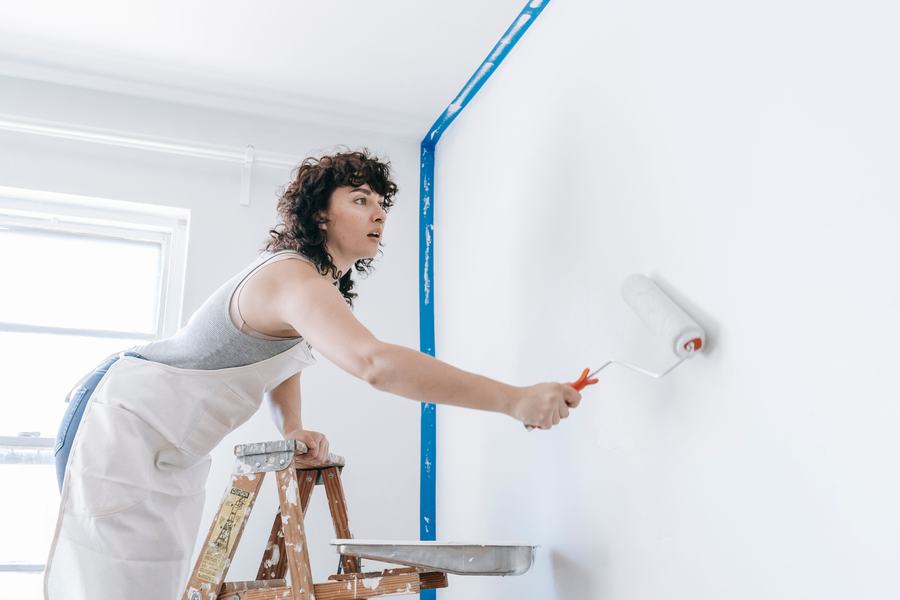 Femme debout sur un escabeau passant un rouleau de peinture sur un mur blanc
